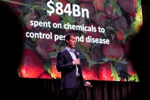 Adam Stager stands on a stage, wearing a blue button-up shirt and blazer and holding a microphone. He stands in front of a projected slideshow which shows the words "$84Bn spent on chemicals to control pests and disease" in white font over a picture of strawberries.