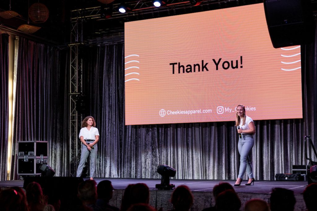 Two women stand on stage, smiling in front of a large projector screen that reads "Thank You!"