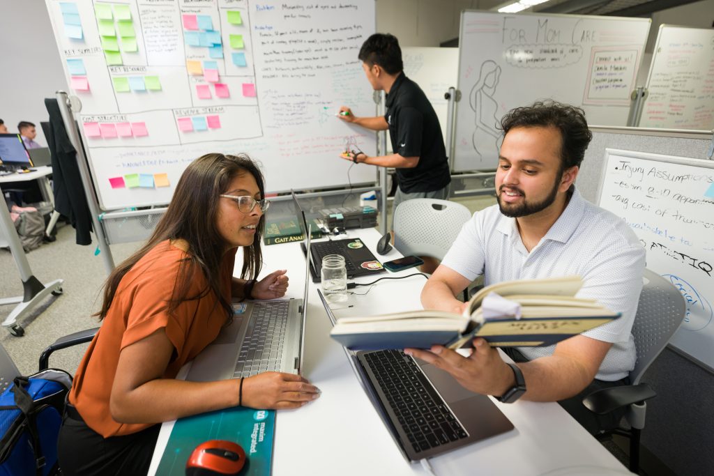Two students sit at a table with a laptop in between them. One student is holding a textbook, and the other is leaning over the table to look at it. In the background, a third student writes on a whiteboard covered in sticky notes.