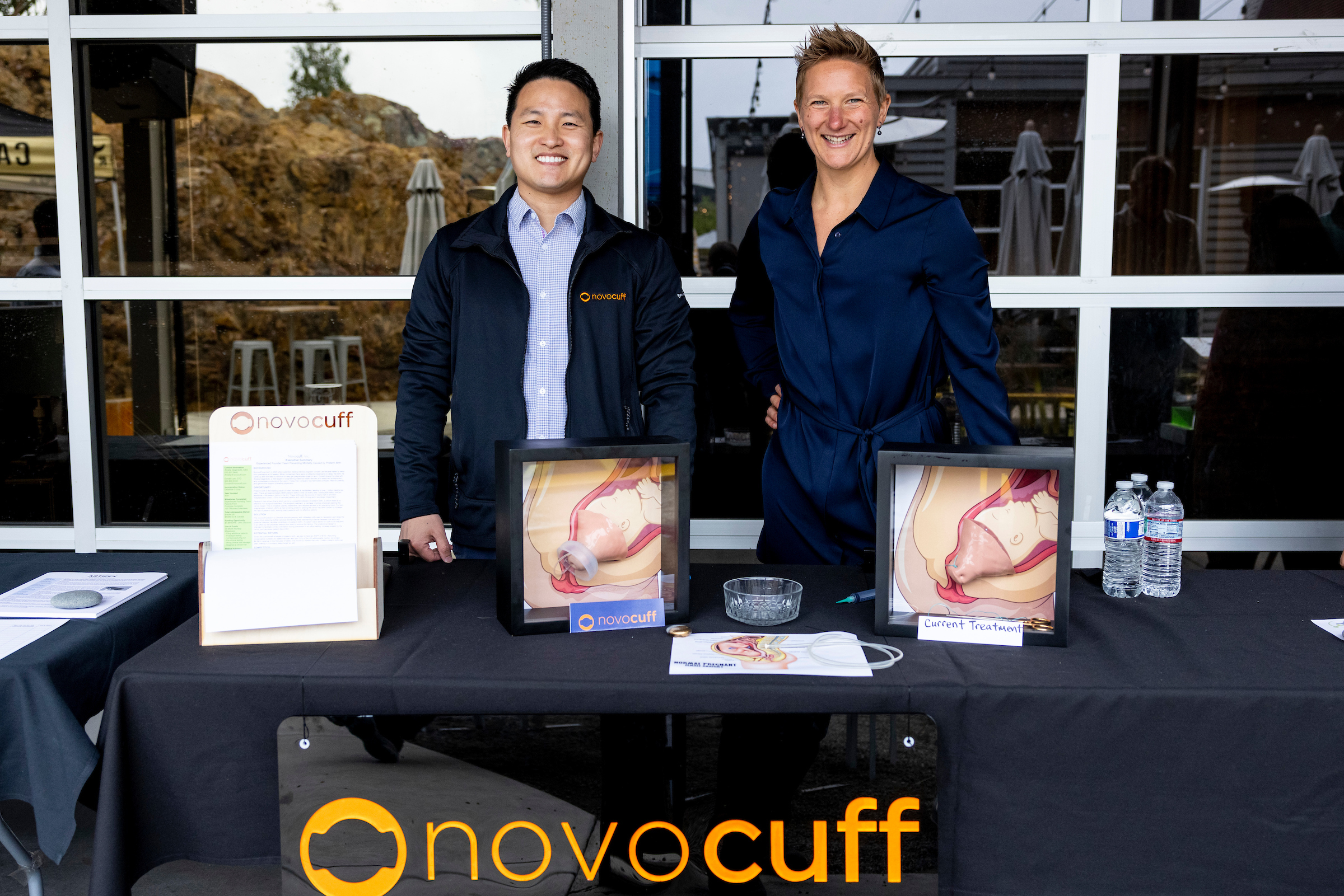Novocuff co-founders Donald Lee (left) and Amelia Defenkolb (right) stand behind a table with images of a fetus in danger of preterm birth with Novocuff's product versus without, as well as their company's logo.
