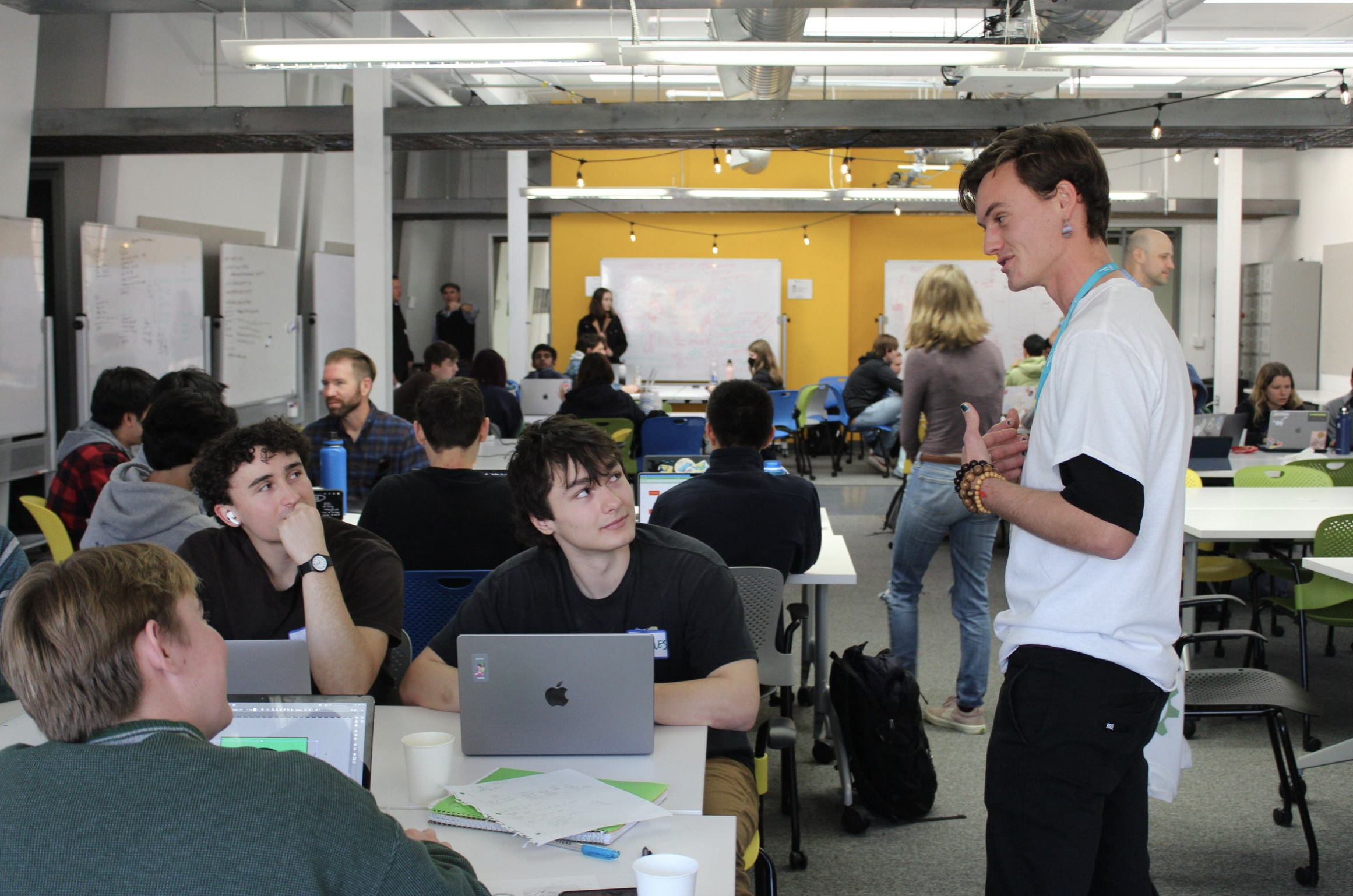 A group of students brainstorm in the CIE HotHouse. One is standing and talking to three other students, seated at a rectangular table with laptops in front of them.