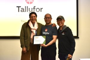 Three people stand in front of a projector screen that reads "Tallyfor." The woman on the left-hand side has her arm around the man in the middle, who is holding a framed paper that reads "Certificate of Graduation." The man on the right-hand side also has his arm around the man in the middle.