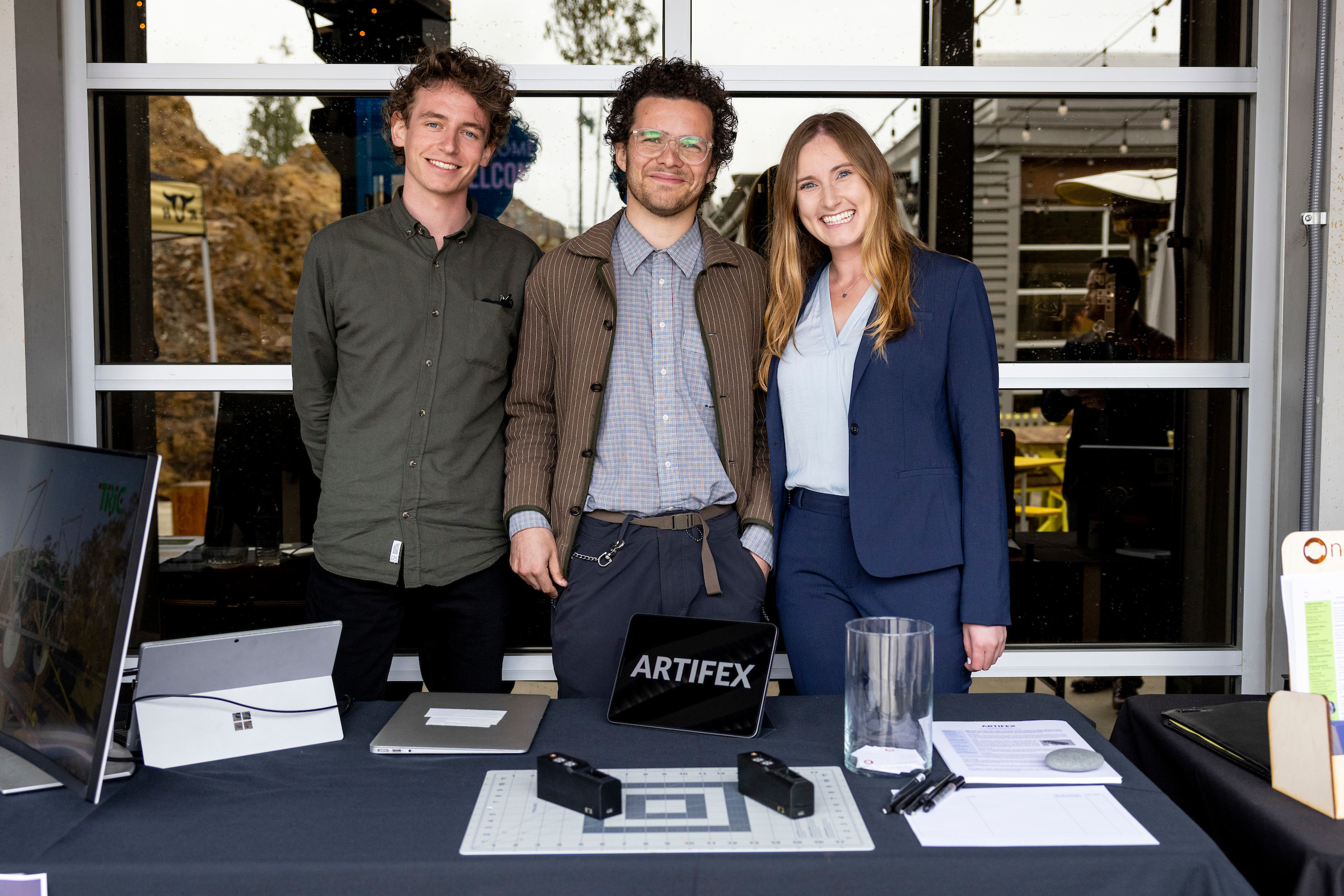 Three people stand behind a table smiling. On the table are blueprint pages and a tablet with the name ARTIFEX displayed on it.