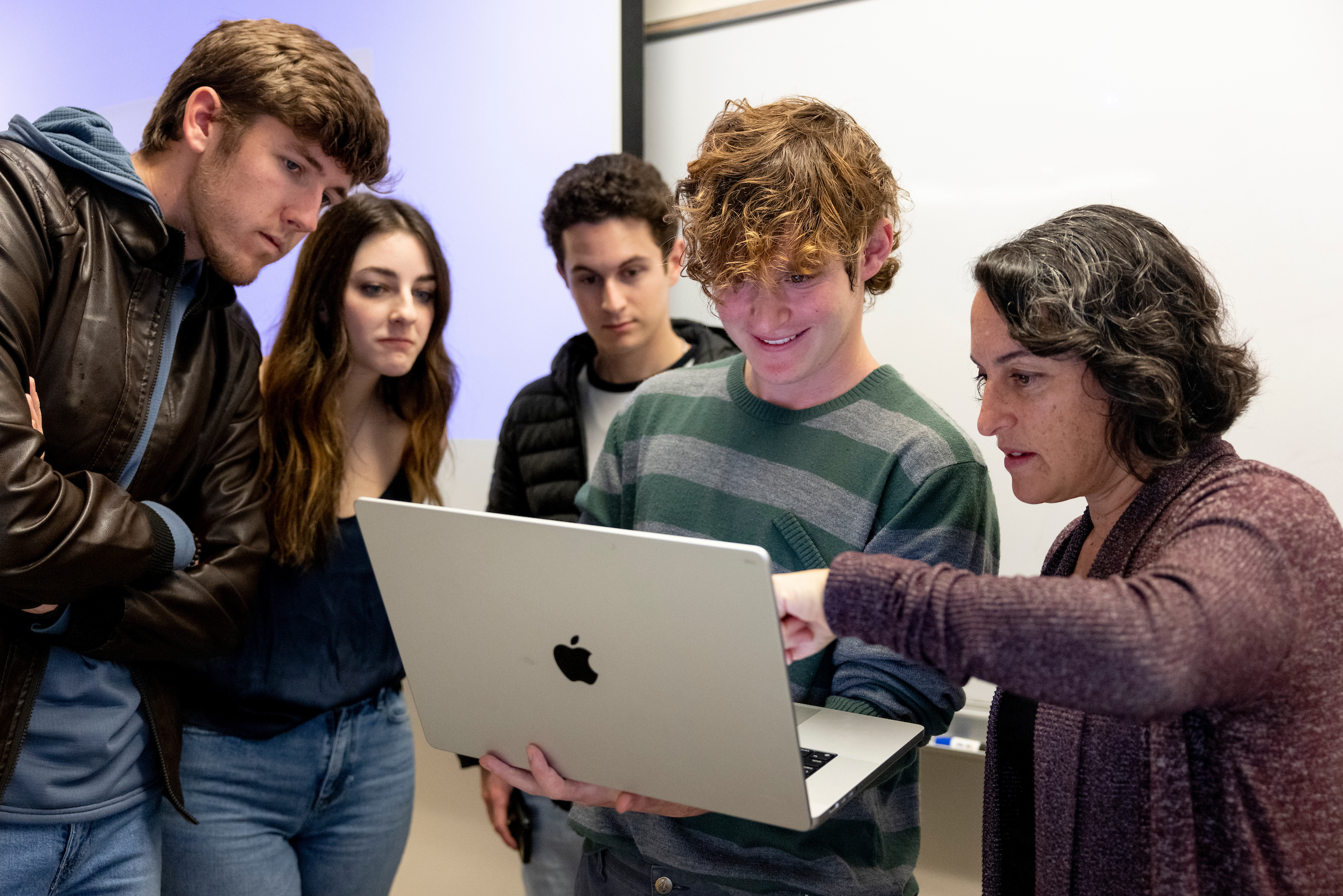 A group of students huddle around a laptop, held by a student in the middle. A professor looks at the laptop and points at the screen.